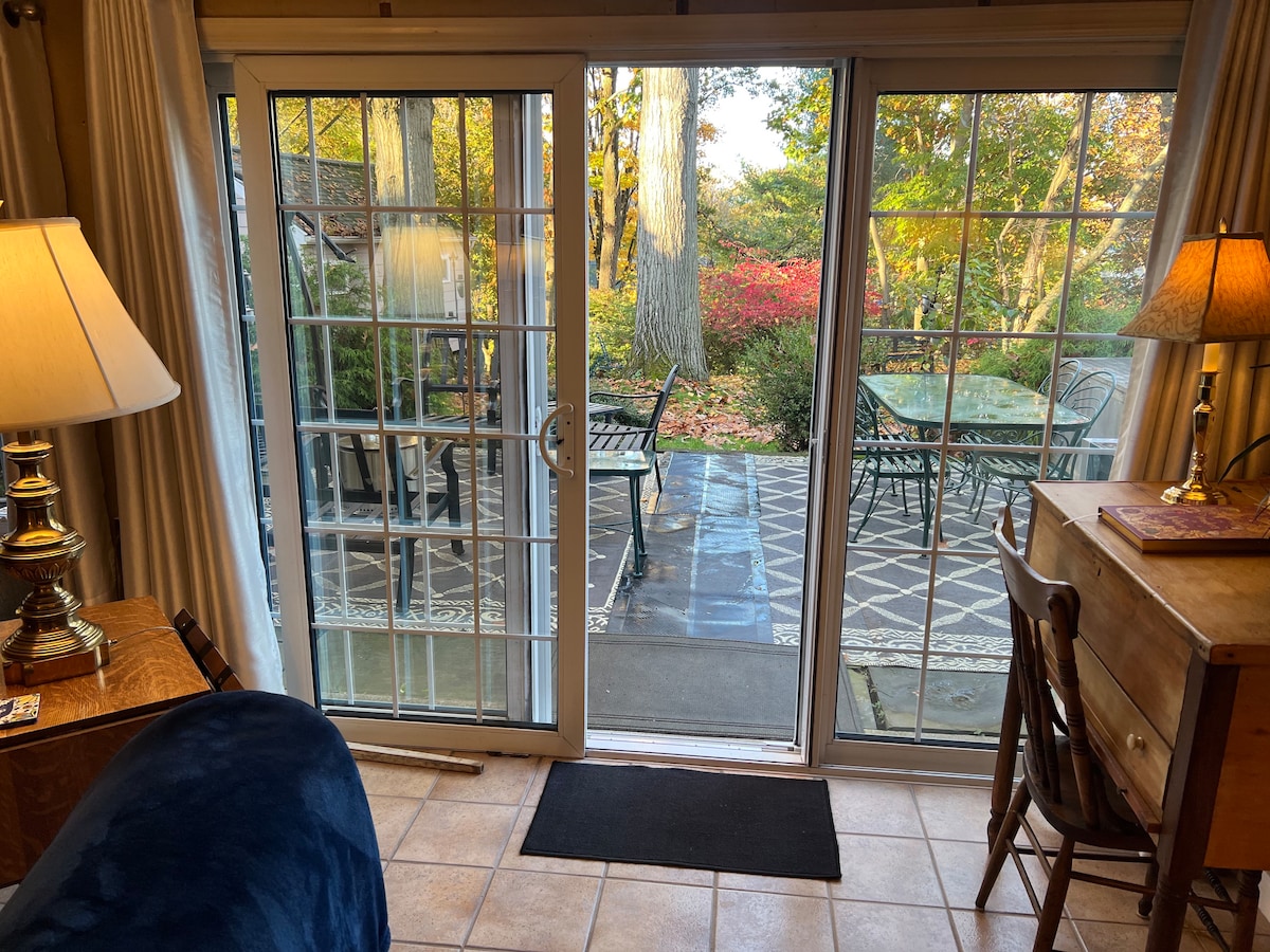 Double glass doors provide a view of a private patio area with outdoor seating, including a table and chairs. The surrounding garden features trees and fall foliage, creating a serene outdoor space. Soft light illuminates the interior area, enhancing the inviting atmosphere.