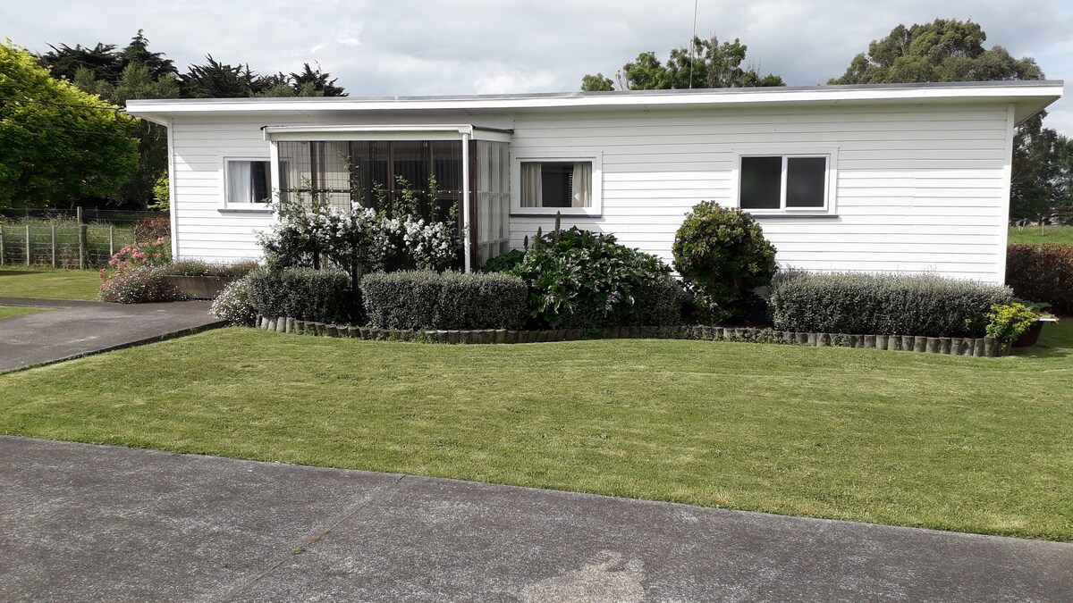 A single-story white building is set within a landscaped garden, featuring various shrubs and flower beds. The entryway is marked by a covered porch with large glass windows. A well-maintained lawn surrounds the structure, with trees and greenery visible in the background.