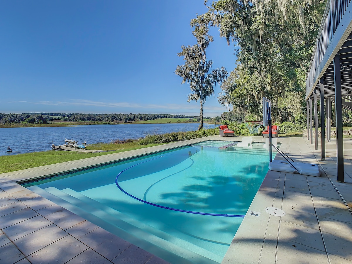 A serene scene features a sparkling pool with a clear view of Dowling Lake in the background. Sunlight reflects off the water, while trees gently sway in the breeze. Comfortable seating areas are available nearby for relaxation and enjoyment of the outdoor space.