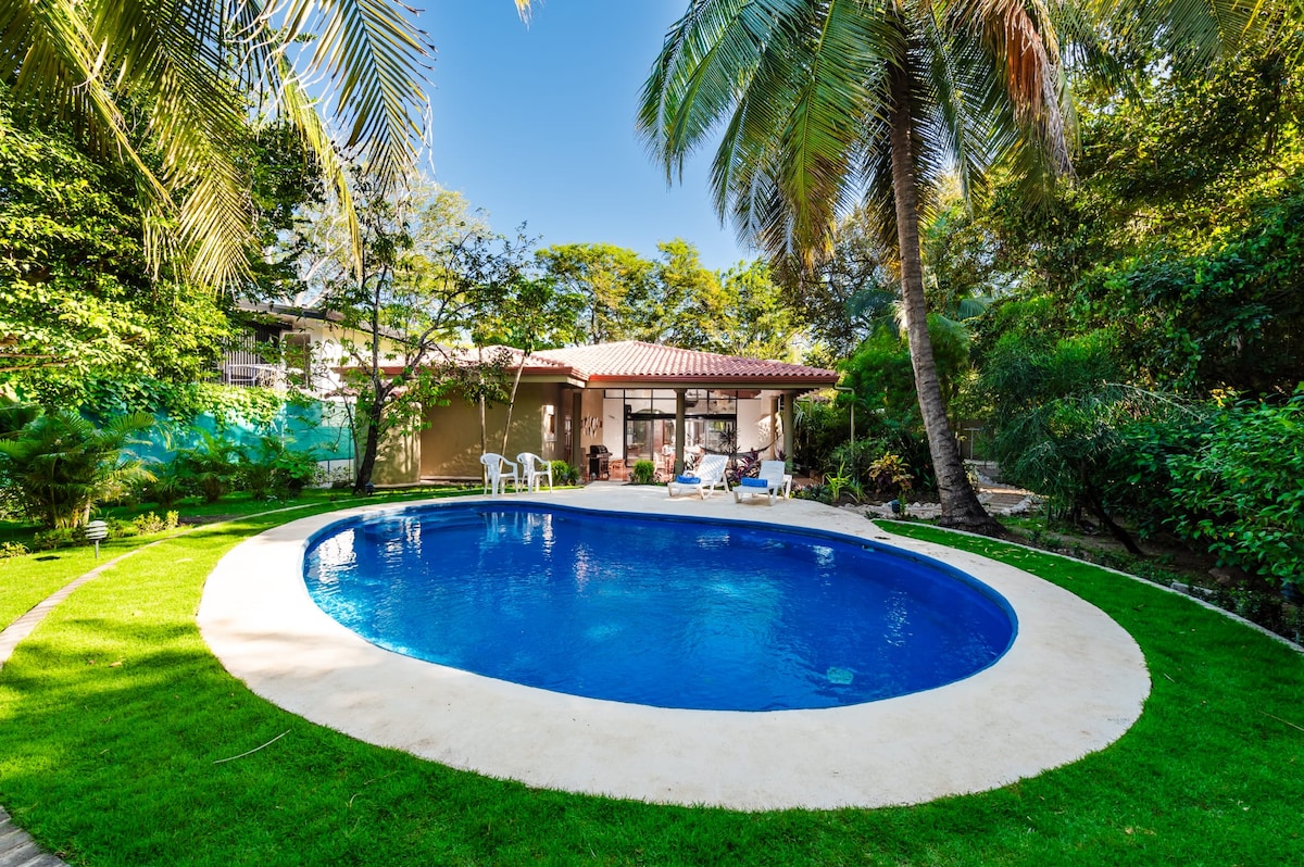 A large, inviting pool is surrounded by a manicured lawn and tropical plants. The house is seen in the background with sliding glass doors that open to the outdoor space. Palm trees provide shade, enhancing the serene environment.
