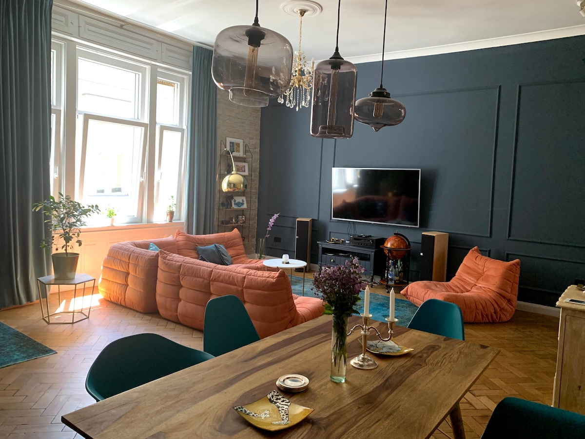 The living area features a spacious layout with large windows allowing natural light to fill the space. Comfortable orange lounge seating is arranged around a low table. A dining table sits in the foreground, complemented by teal chairs, while a television and entertainment system are positioned against a dark accent wall.