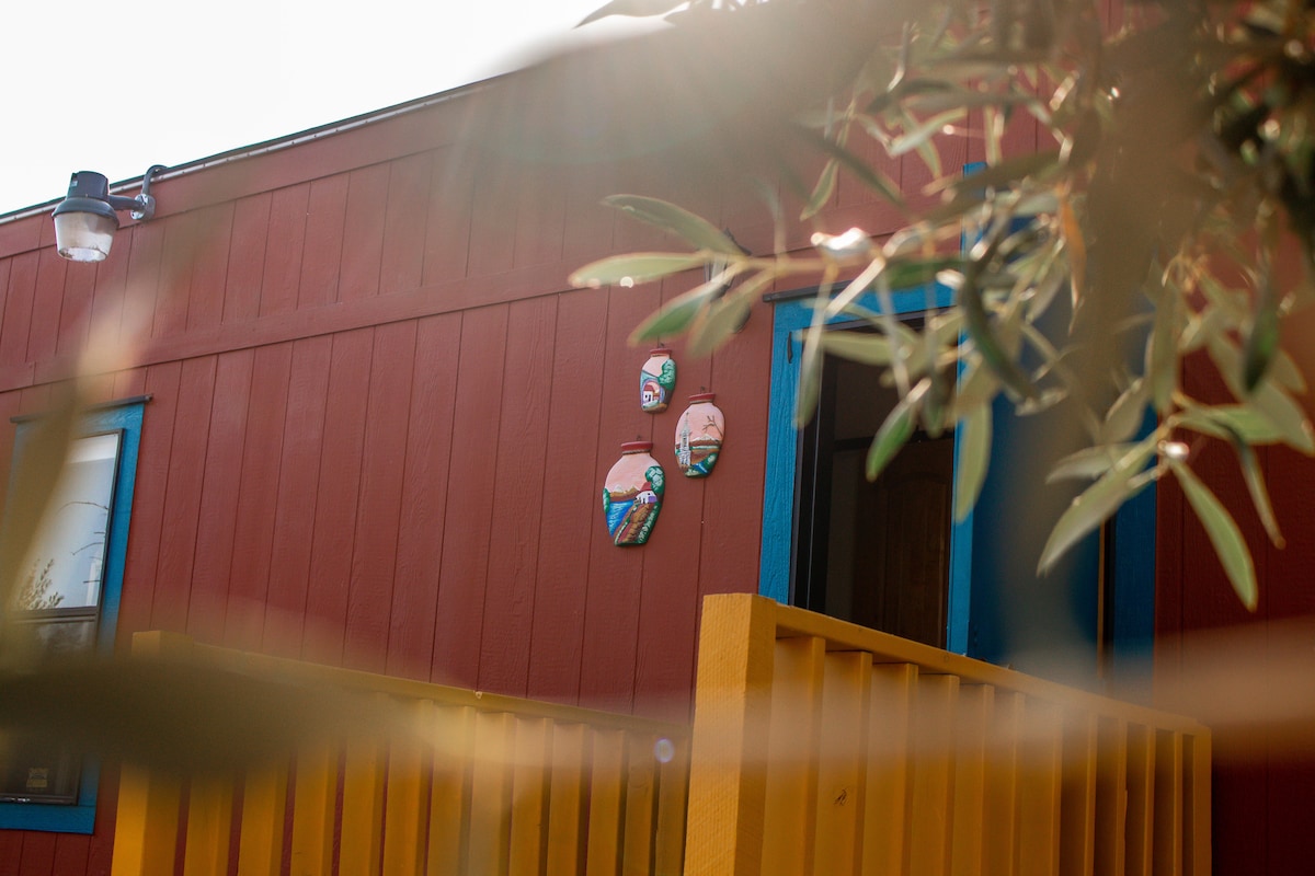 The exterior of a colorful building is visible, featuring a red façade with bright blue trim. Three decorative wall pieces can be seen, adding a touch of artistry. Olive leaves frame the image, indicating the surrounding greenery.