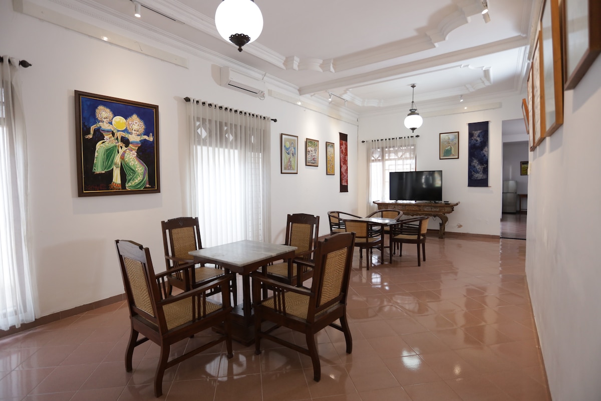 A spacious dining area is showcased with four wooden tables and eight wooden chairs, arranged for comfort. Natural light filters through sheer white curtains, illuminating the walls adorned with vibrant artwork. Soft lighting from ceiling fixtures enhances the welcoming ambiance, ensuring a cozy space for gatherings.