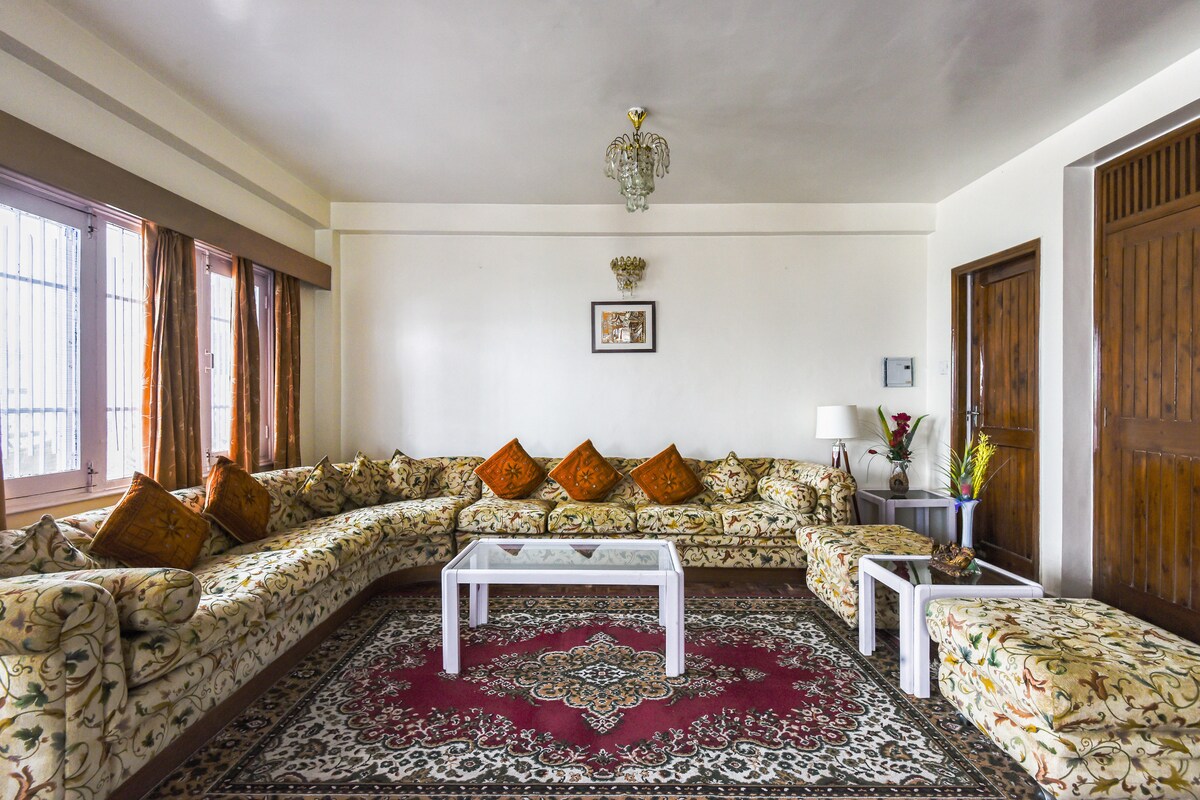 A spacious living area features a large floral-patterned sectional sofa adorned with orange cushions. A white coffee table is placed on a decorative rug, with large windows allowing natural light to brighten the room. A lamp and plants are included for added decor.