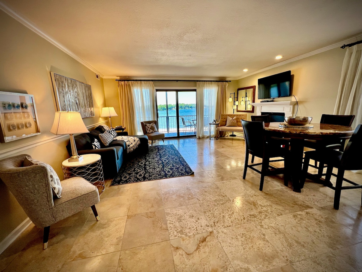 An open-concept living area displays comfortable furnishings and a dining table for four. Natural light fills the space through large glass doors that lead to a private balcony overlooking the water. Neutral tones and decorative accents create a serene ambiance.