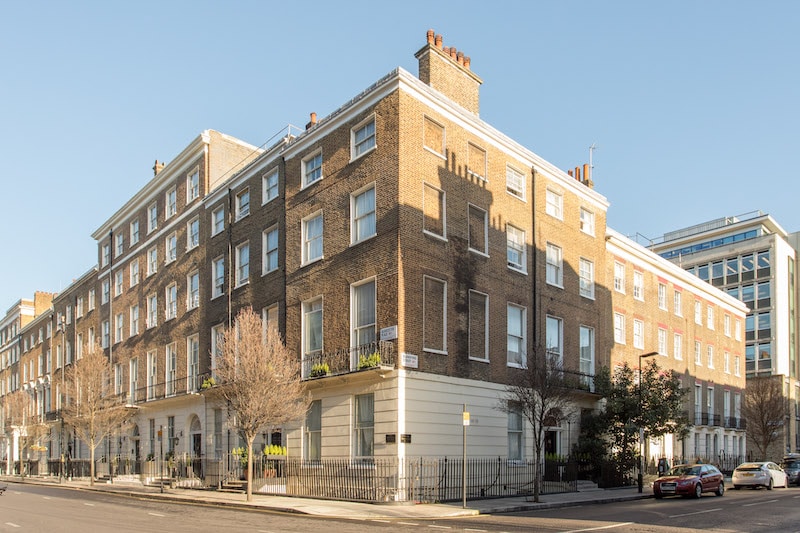 A charming corner view of the Residence Apartments building is displayed. The structure features a classic brick façade with multiple windows, some adorned with window boxes. The ground level showcases a welcoming entrance, complemented by greenery along the pathway.