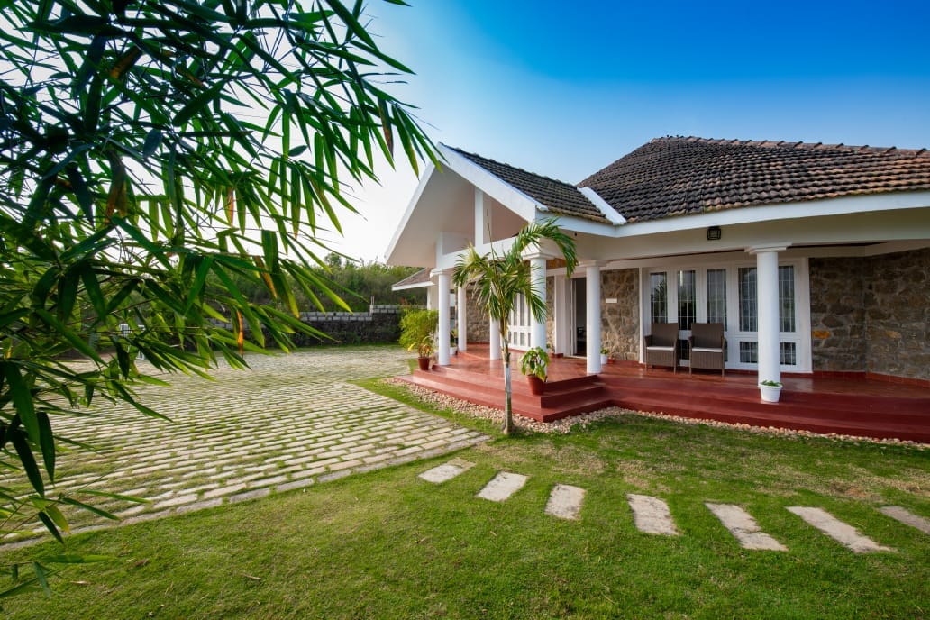The exterior of Serenity Villa is showcased, featuring a stone façade complemented by wooden accents. A spacious front yard is visible, bordered by a path of stones leading towards the entrance. Lush greenery surrounds the property, providing a serene atmosphere.
