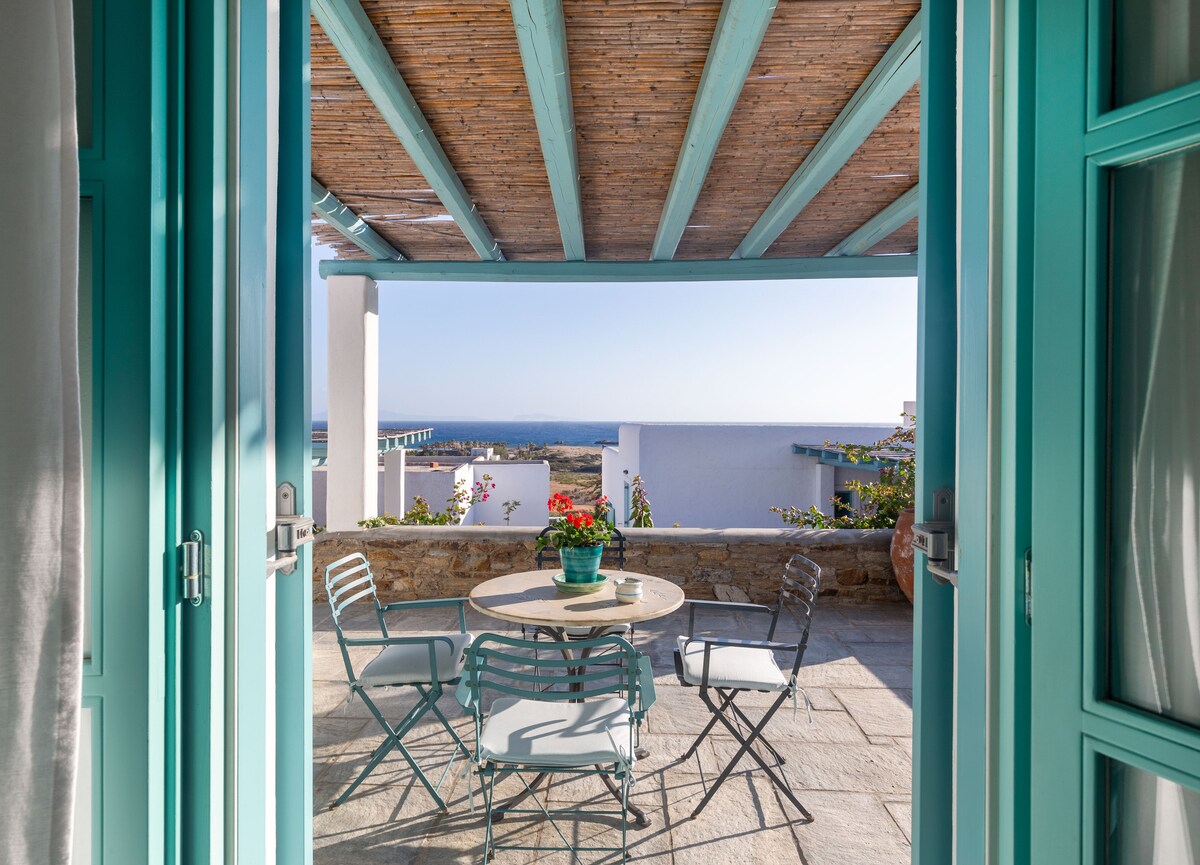 Double doors open to a shaded terrace featuring a round table surrounded by four metal chairs. A potted plant sits at the center, with a panoramic view of the sea visible beyond the stone wall. The wooden ceiling adds a rustic touch.