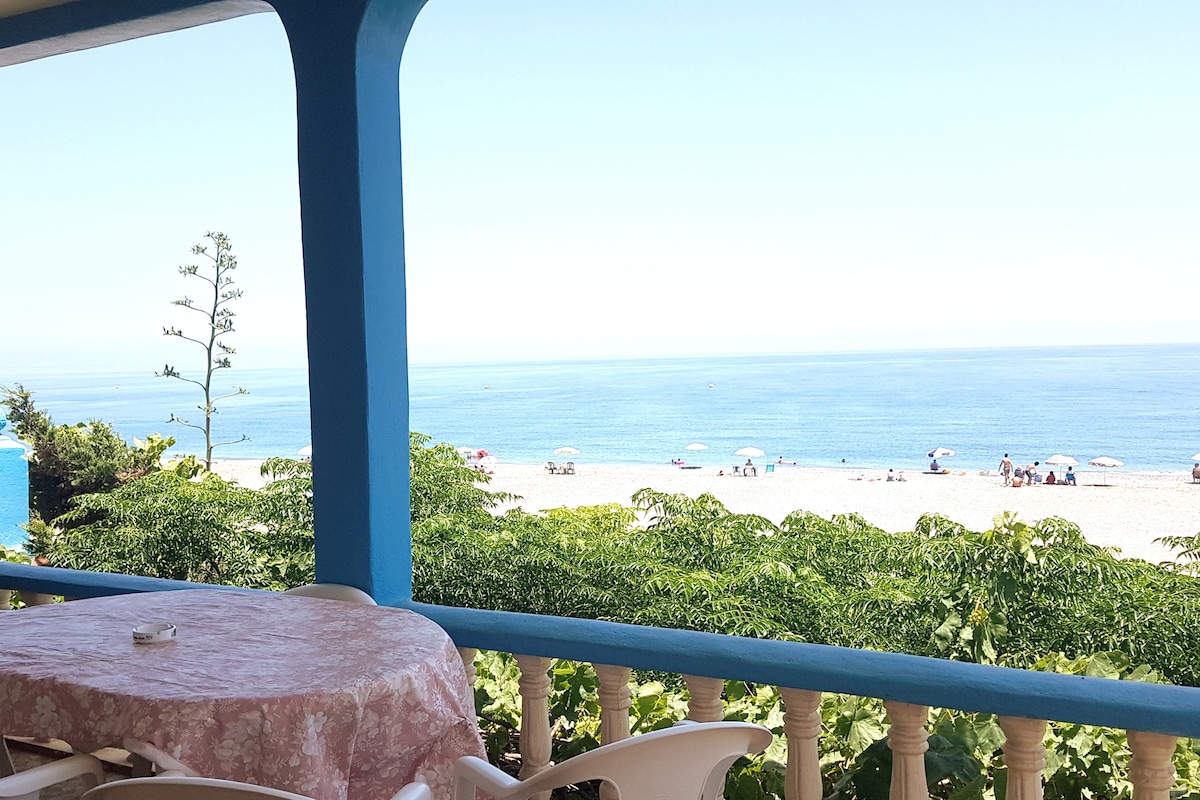 The image displays a spacious terrace with a round table, offering a clear view of the beach and the Mediterranean Sea. Sunbathers are visible on the sandy shore, with colorful umbrellas dotting the landscape. Lush greenery frames the scene, adding a natural touch.
