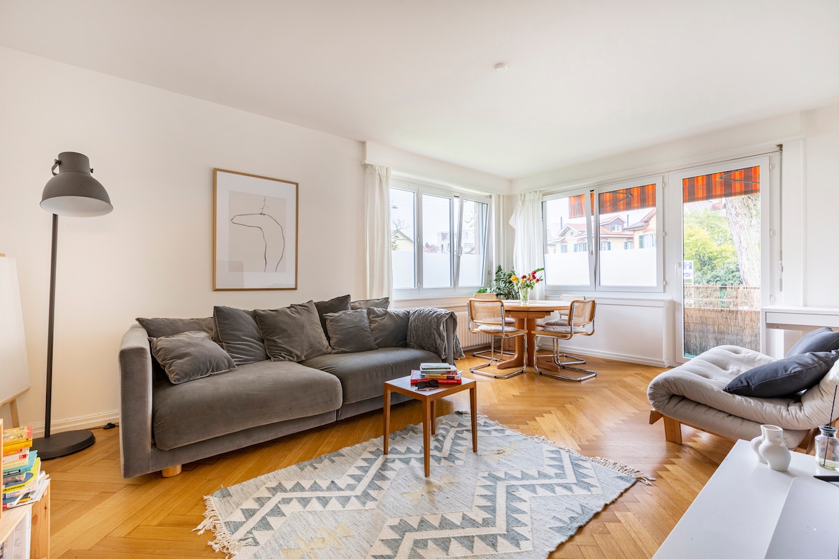 A bright living area features large windows allowing natural light to fill the space. A spacious gray sectional sofa is positioned around a wooden coffee table, complemented by a patterned area rug. A dining table for four, surrounded by light-colored chairs, is also visible.