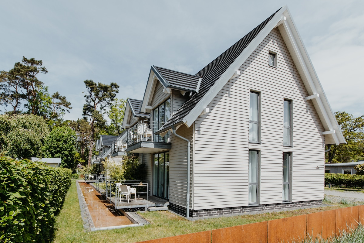 The exterior of a modern two-story building is presented, featuring a light-colored wooden façade. Large windows reflect natural light, while balconies provide outdoor seating. Surrounding greenery enhances the serene environment, contributing to the inviting appearance of the property.