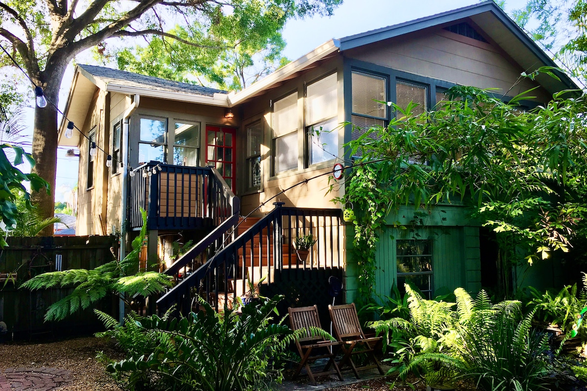 A charming two-story bungalow surrounded by lush greenery and outdoor seating. The entrance features a set of steps leading to a wooden porch, framed by large windows that invite natural light. The yard includes various plants, enhancing the inviting atmosphere.