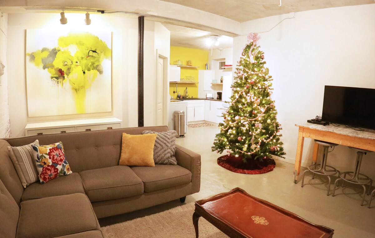 An industrial modern living space showcases a large gray sectional sofa adorned with colorful pillows. A coffee table with ornate decoration is positioned in front of the sofa. The room features a Christmas tree decorated with lights and ornaments, and a bright artwork hangs on the wall.