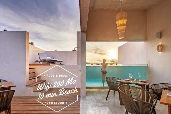 A serene rooftop space features a swimming pool with a view and a cozy dining area. Sunlight reflects off the water, highlighting a figure enjoying the tranquil setting. Elegant furnishings include lounge chairs and dining tables, contributing to a relaxing atmosphere surrounded by nature.