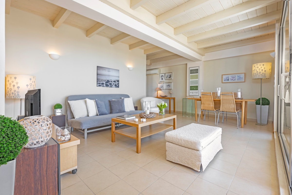A spacious living area is illuminated by abundant natural light, featuring a comfortable sofa, a wooden coffee table, and a soft ottoman. A dining table with woven chairs is positioned in the background, complemented by decorative lamps and a touch of greenery in stylish pots.
