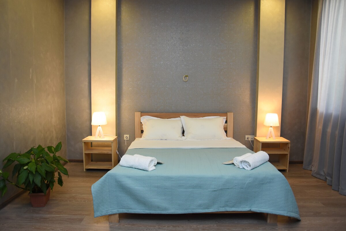 A spacious bed is presented with light-colored linens, complemented by neatly arranged towels at its foot. Soft lighting from bedside lamps illuminates the room, which features a muted wall design and a potted plant in the corner, creating a calming atmosphere.