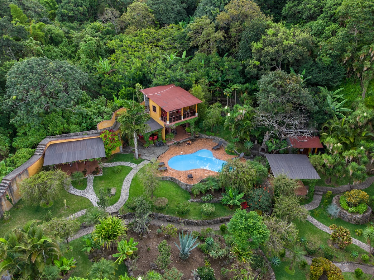 An aerial view of Villa Eggedal showcases its vibrant gardens and inviting pool. Lush greenery surrounds a central swimming area with a softly curving shape, while pathways meander through the well-maintained landscape, leading to colorful flowers and shaded seating areas.