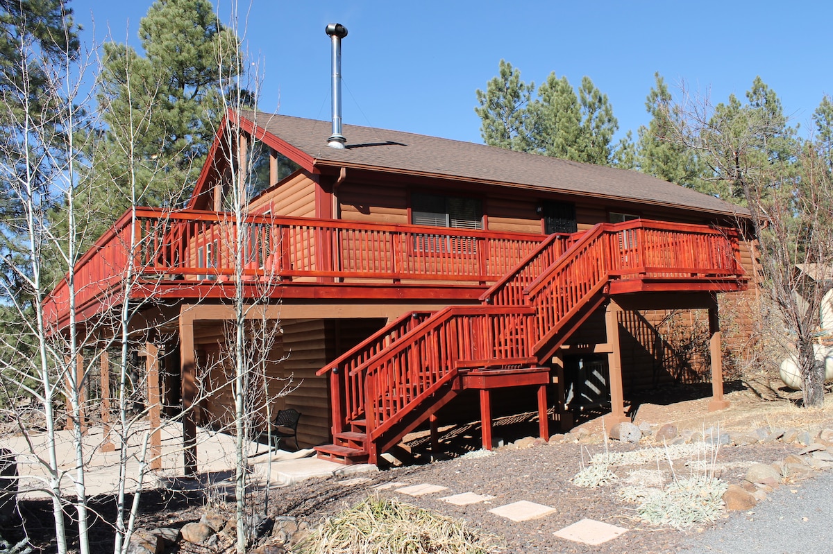 A spacious log cabin is set among tall ponderosa pines, featuring a large red wrap-around deck and a set of stairs leading up to the entrance. The structure showcases a warm wooden exterior with a chimney, inviting outdoor spaces that encourage relaxation and enjoyment.