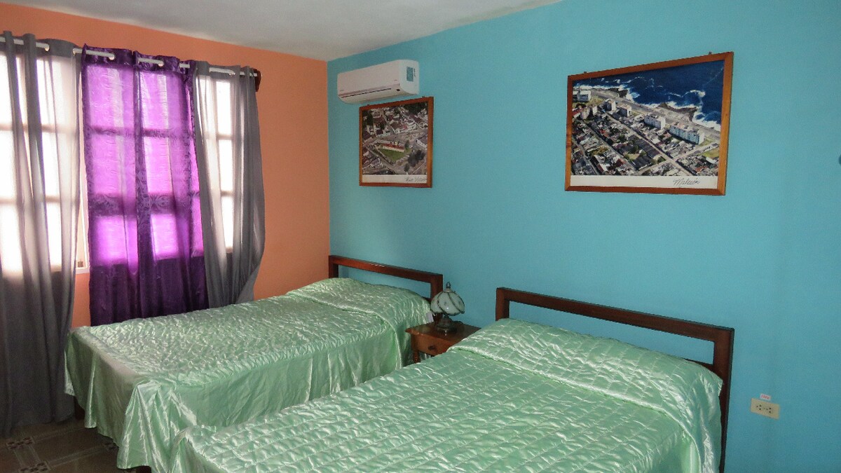 A bright room features two beds with green bedspreads. Light floods in through windows draped with sheer purple curtains. Cool-toned walls are complemented by framed artwork depicting coastal scenes, adding visual interest to the space.