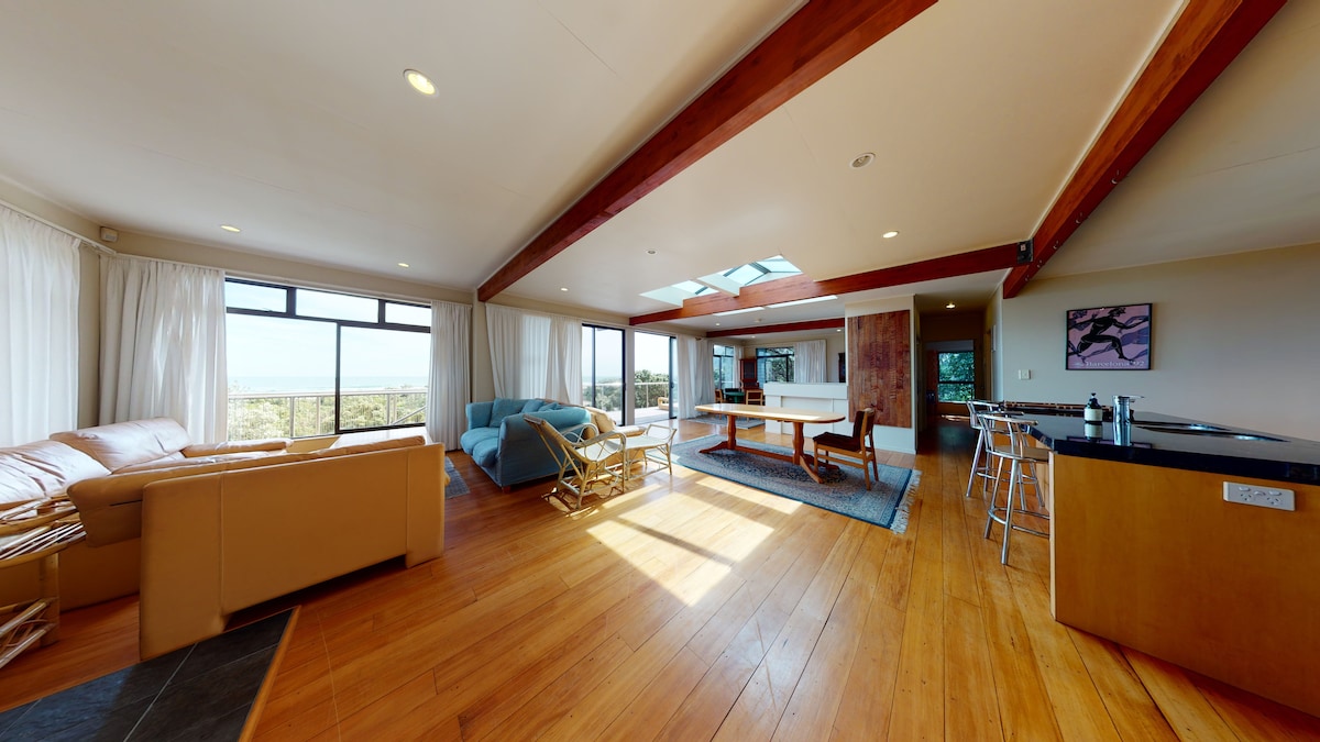 An open-plan living area is highlighted by large windows allowing natural light to fill the space. A blue sofa and a light brown leather couch provide comfortable seating. A wooden dining table is positioned near the kitchen, featuring modern bar stools along the counter.