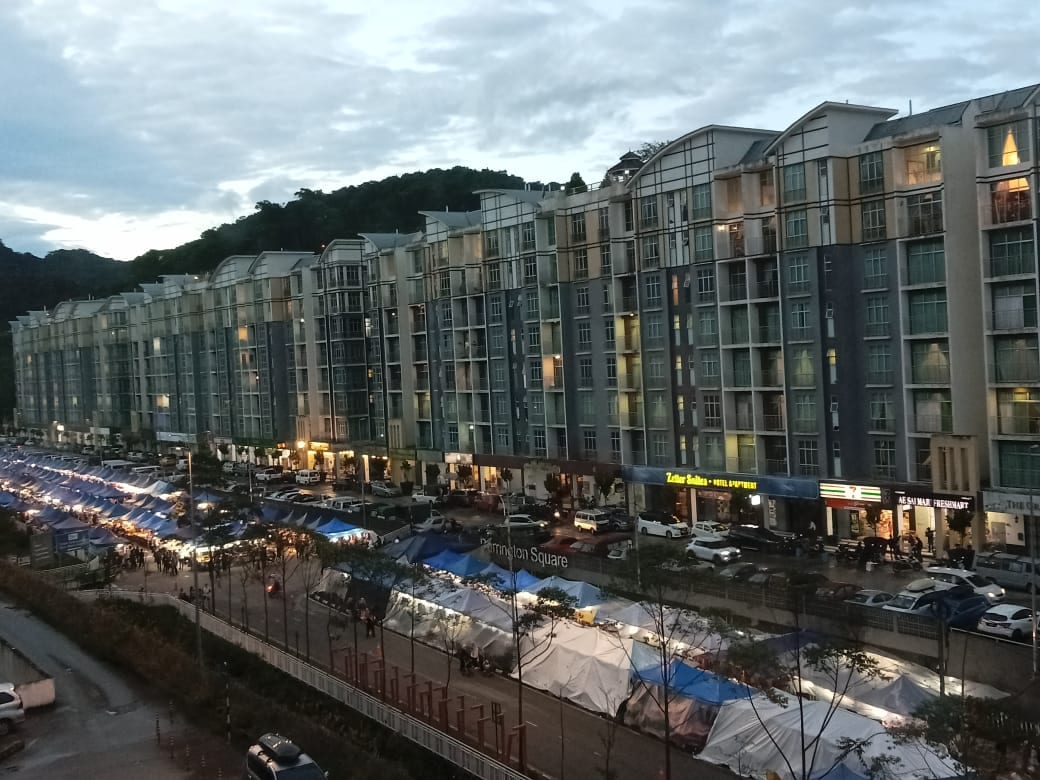 A view of the street below reveals a vibrant night market with numerous stalls covered by blue and white tents. The surrounding apartment buildings are illuminated, showcasing their modern architecture against a backdrop of a cloudy evening sky and the hillside.