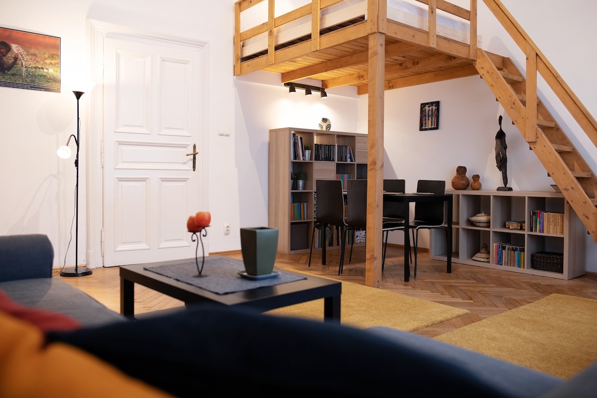 A cozy living area features a comfortable sofa and a coffee table, with a decorative bowl of fruit. The dining space includes a wooden table surrounded by four chairs, situated next to bookshelves filled with books. A lofted sleeping area is accessible by a wooden staircase.