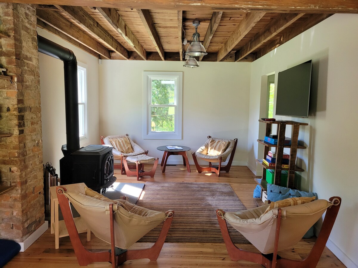 A cozy living area is highlighted by two fabric chairs and a wooden coffee table, with a seasonal wood stove placed against a brick feature wall. Natural light filters through two windows, illuminating the room's warm wood accents and simple decor.