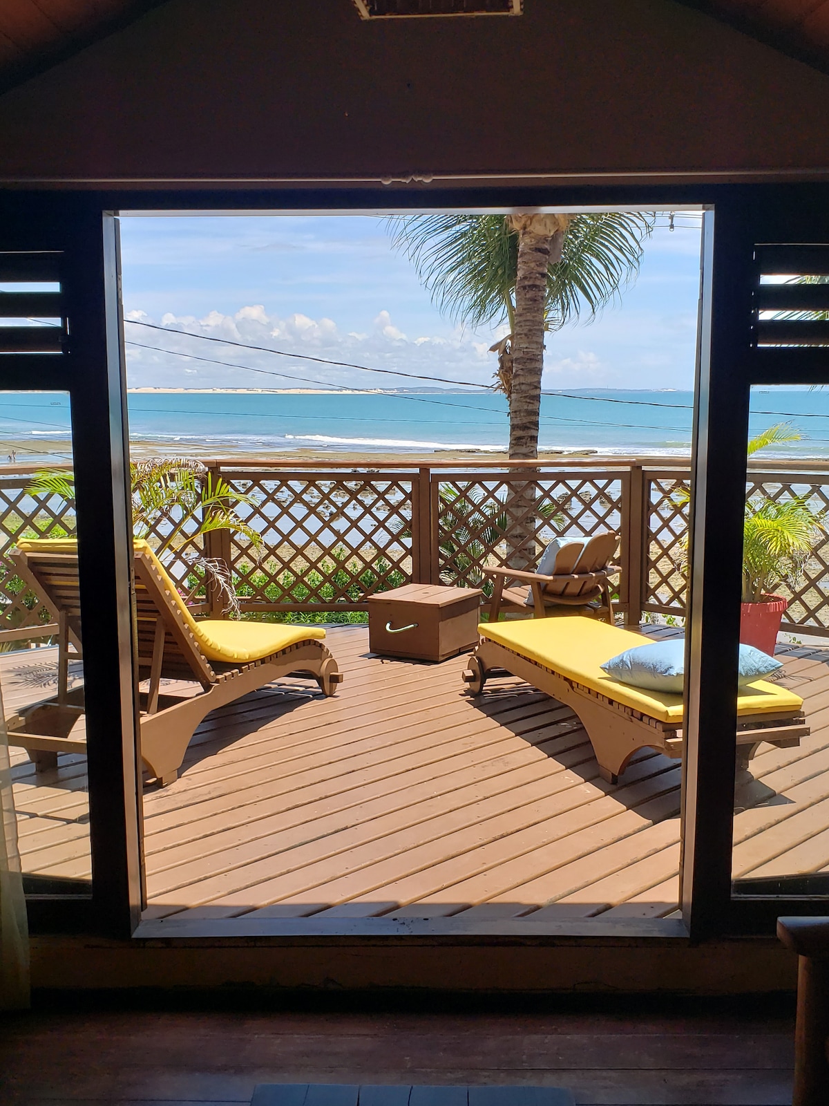 A terrace is showcased with two yellow lounge chairs facing the ocean, surrounded by tropical greenery. The view beyond reveals the turquoise sea and palm trees, inviting natural light into the space. Wooden deck flooring enhances the connection with the outdoor environment.