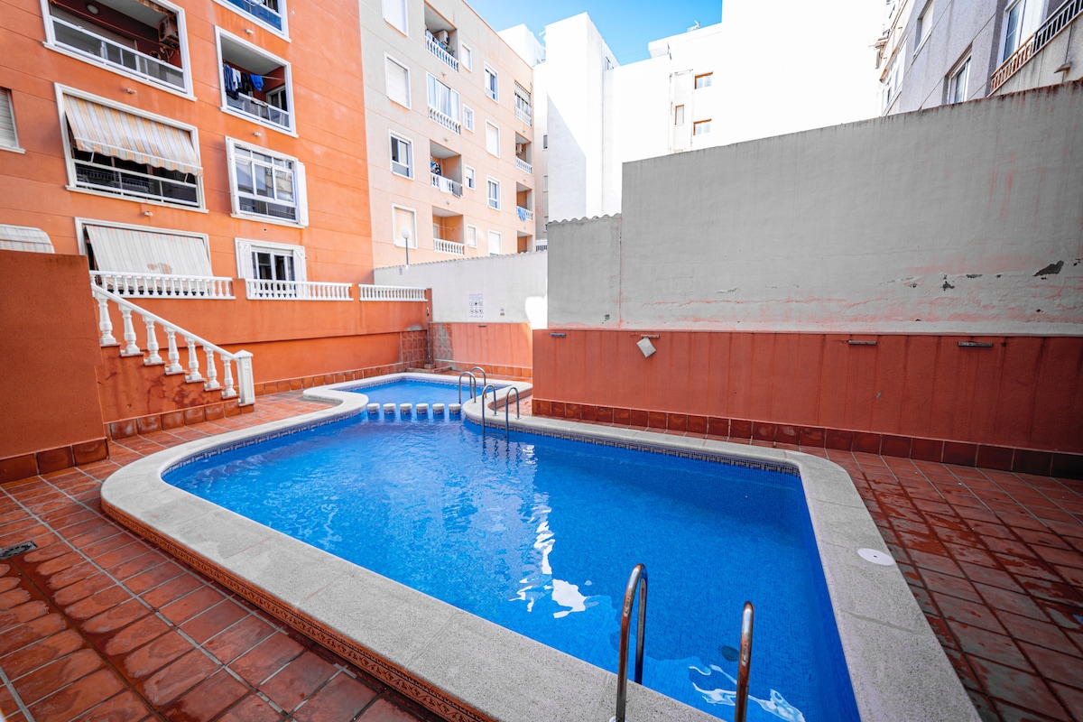 A shared outdoor pool is visible, surrounded by a smooth patio with terracotta tiles. Steps lead into the water, which is a calming blue. Adjacent buildings provide a sense of enclosure, while sunlight highlights the pool's inviting surface.