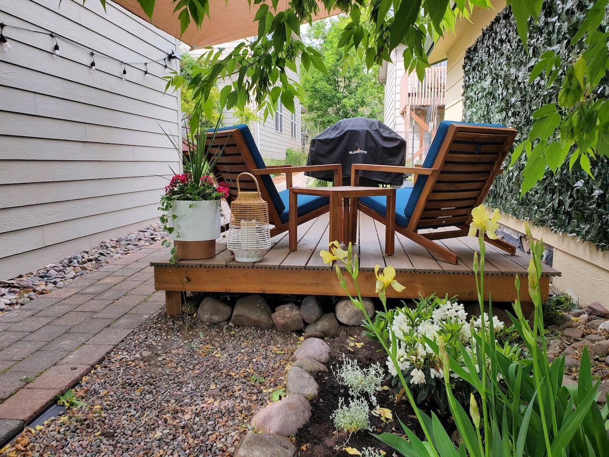 A private deck is shown surrounded by greenery and colorful flowers. Two wooden chairs are positioned around a small table, with a gas grill in the background. Planters with vibrant blooms and a wicker basket add charm to the outdoor space.