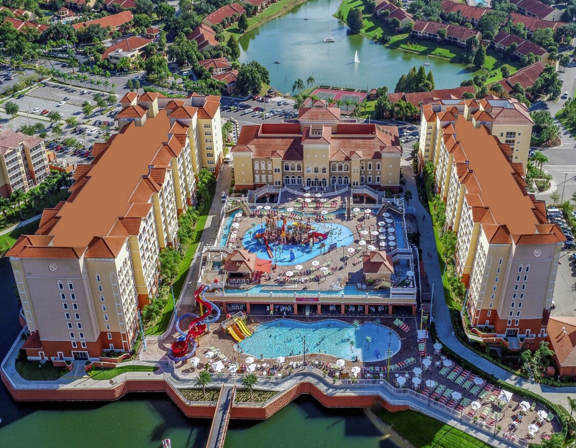 An aerial view showcases the resort's expansive layout, featuring multiple buildings surrounding a large water park area. Colorful water slides and pools are visible, with lounge chairs arranged around the outdoor facilities. Lush greenery and a lake complement the vibrant setting.