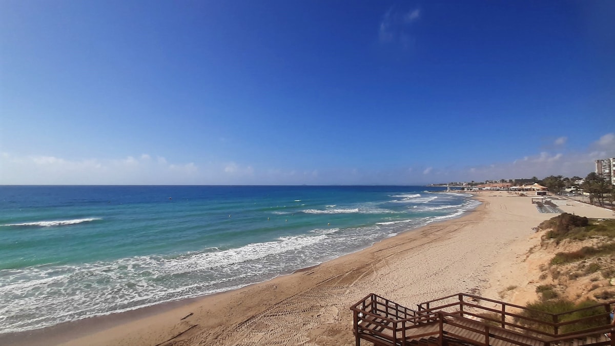 A wide view of a sandy beach with gentle waves lapping at the shore. The horizon is filled with a clear blue sky scattered with soft clouds, while the sparkling water showcases shades of blue and green. Beachside structures are visible in the distance.