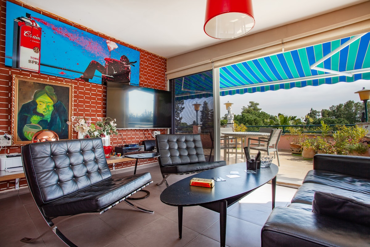 The living area showcases modern black lounge chairs arranged around a sleek, oval coffee table. A large glass door opens to the terrace, allowing natural light and views of greenery. Colorful artwork adorns the wall, adding a vibrant touch to the relaxed environment.