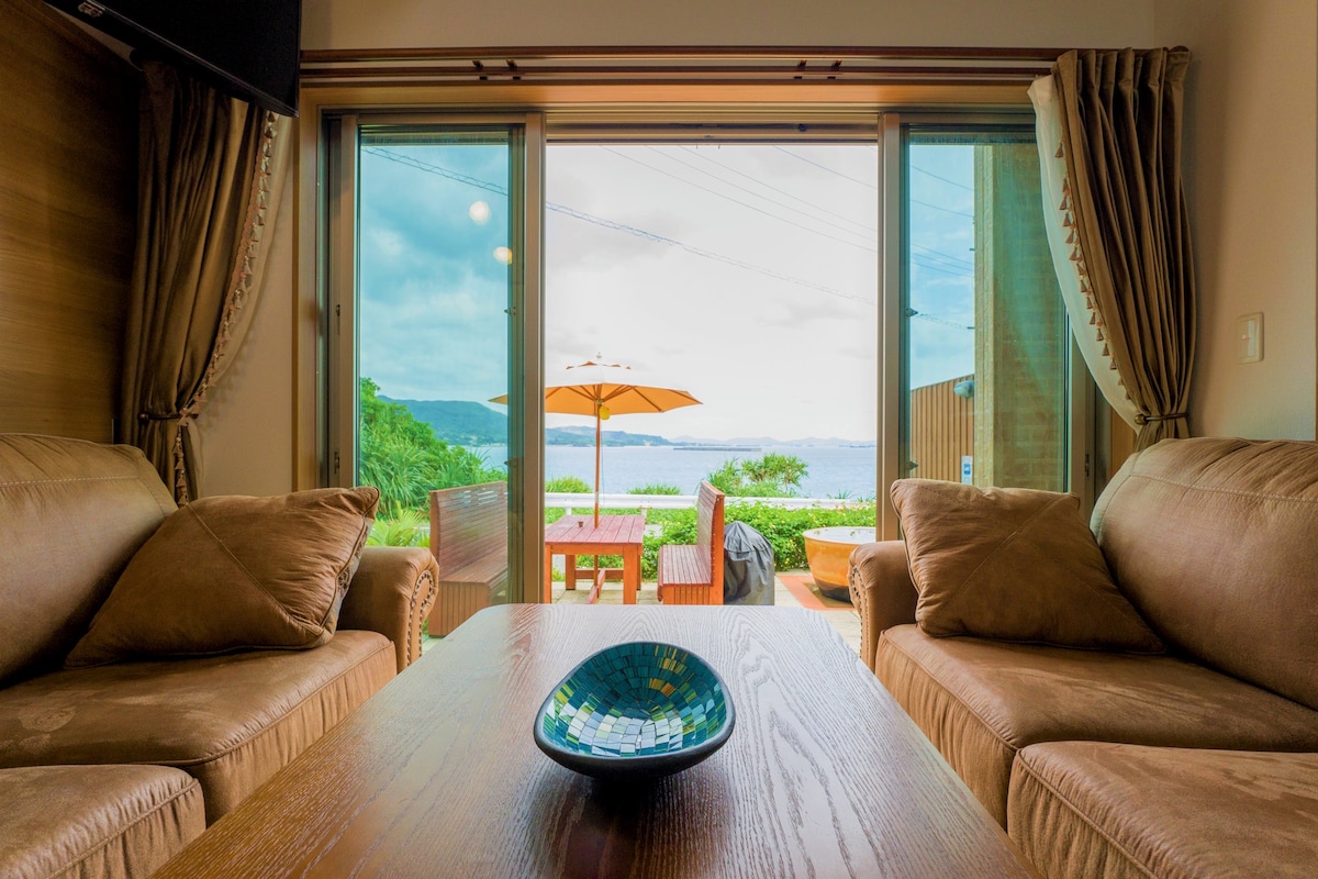 A living room area is presented with two plush sofas facing a wooden table. Large sliding glass doors open to a terrace with a sea view, featuring an umbrella-shaded dining table. Soft natural light fills the room, accentuating the warm colors of the furnishings.