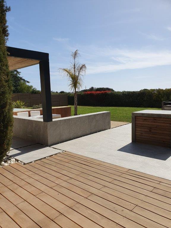An outdoor space is depicted featuring a wooden deck leading to a modern stone fixture. Tropical plants and a well-maintained lawn are visible in the background, with clear blue skies enhancing the light atmosphere.