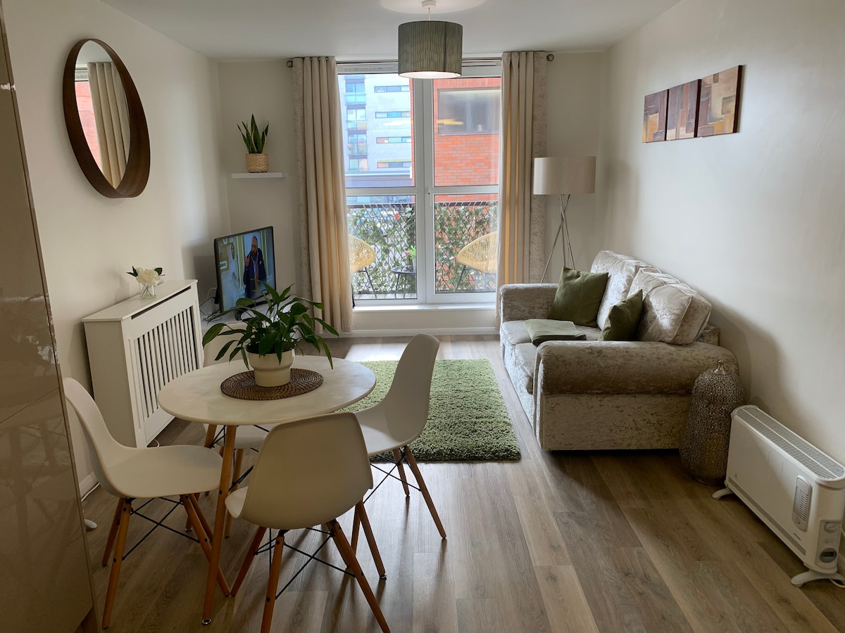 The living area features a circular dining table surrounded by four chairs and a cozy sofa. A potted plant adds a touch of greenery. Natural light washes in through the large window, which is partially dressed with light curtains, enhancing the space's modern feel.