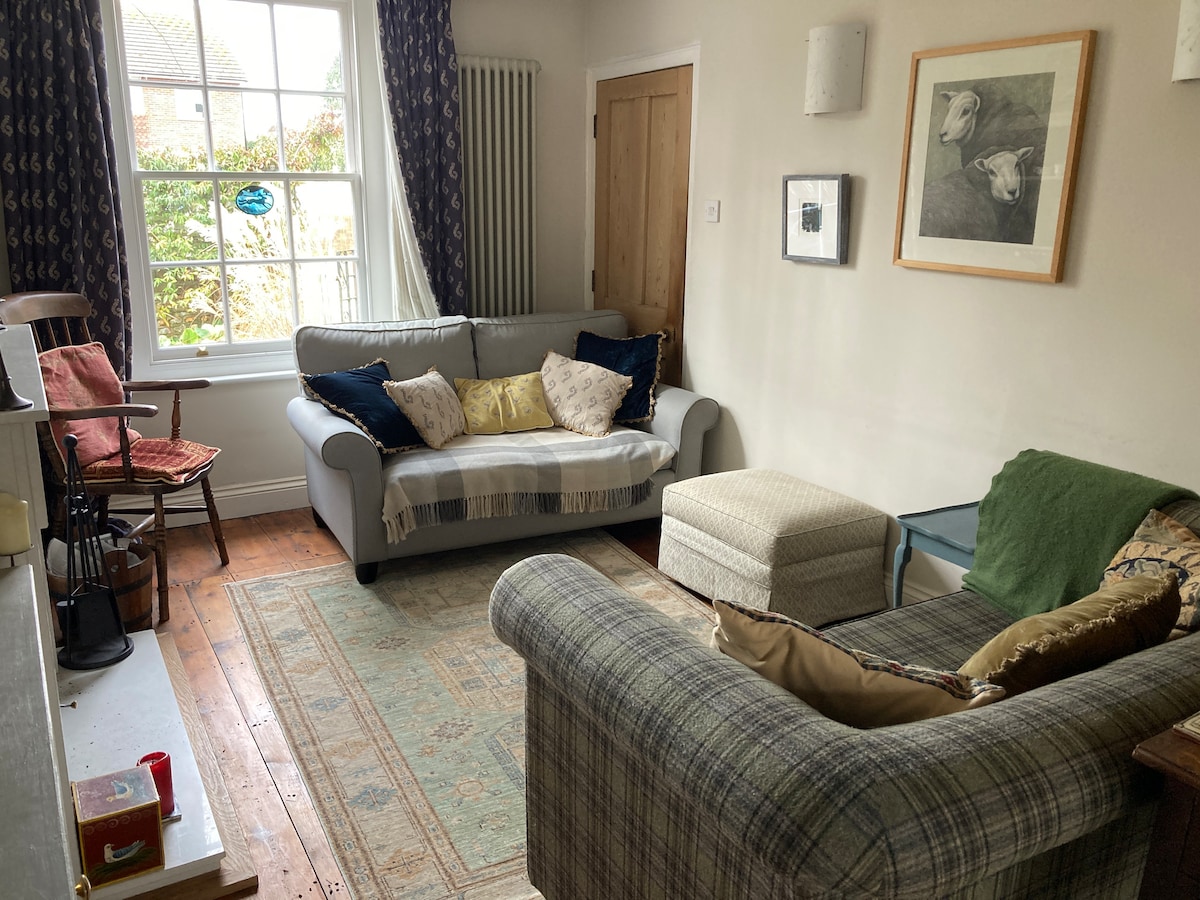 A cozy living area features a mix of seating options, including a plaid sofa and a light grey couch adorned with decorative pillows. Natural light filters through large windows, illuminating a warm wooden floor and a subtle area rug.