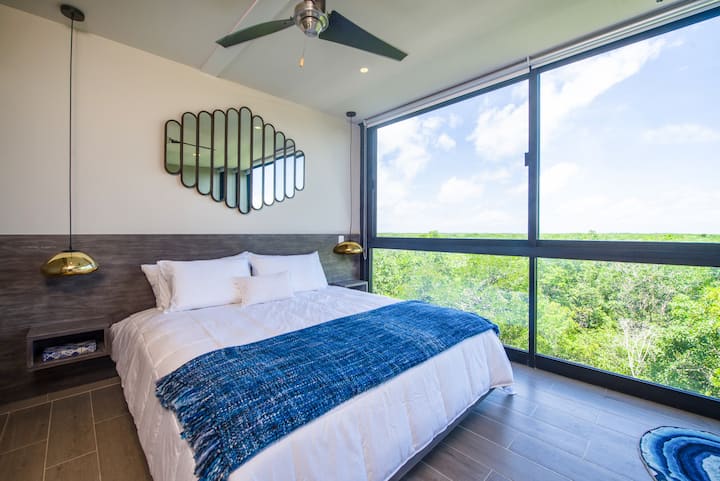 Guest bedroom with king bed, blackout shades, and floor-to-ceiling windows with views of the Mayan Ruins and Tulum National Park.