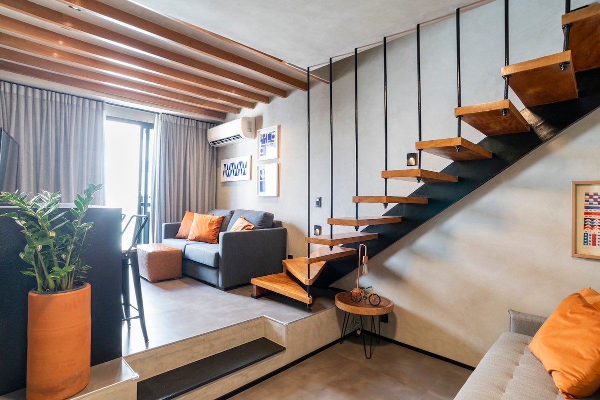 The living area features a modern design with two sofas, one being a comfortable sofa bed. A distinctive wooden staircase leads to the upper level. Large windows allow natural light to fill the space, complemented by tasteful artwork and a potted plant.