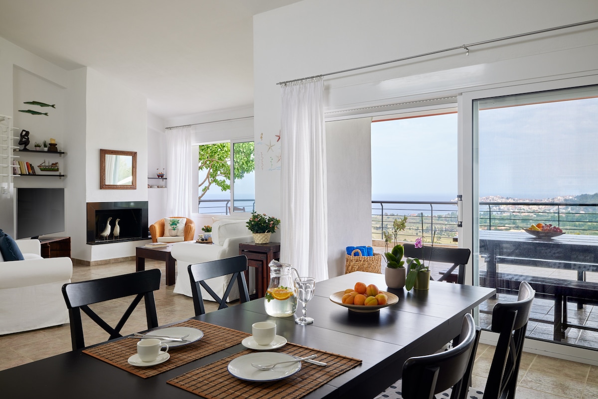 A spacious living and dining area is on display, featuring large glass doors that provide a view of the sea and surrounding landscape. A modern dining table is set with dishes and a bowl of fruits, while comfortable seating is arranged near a fireplace.