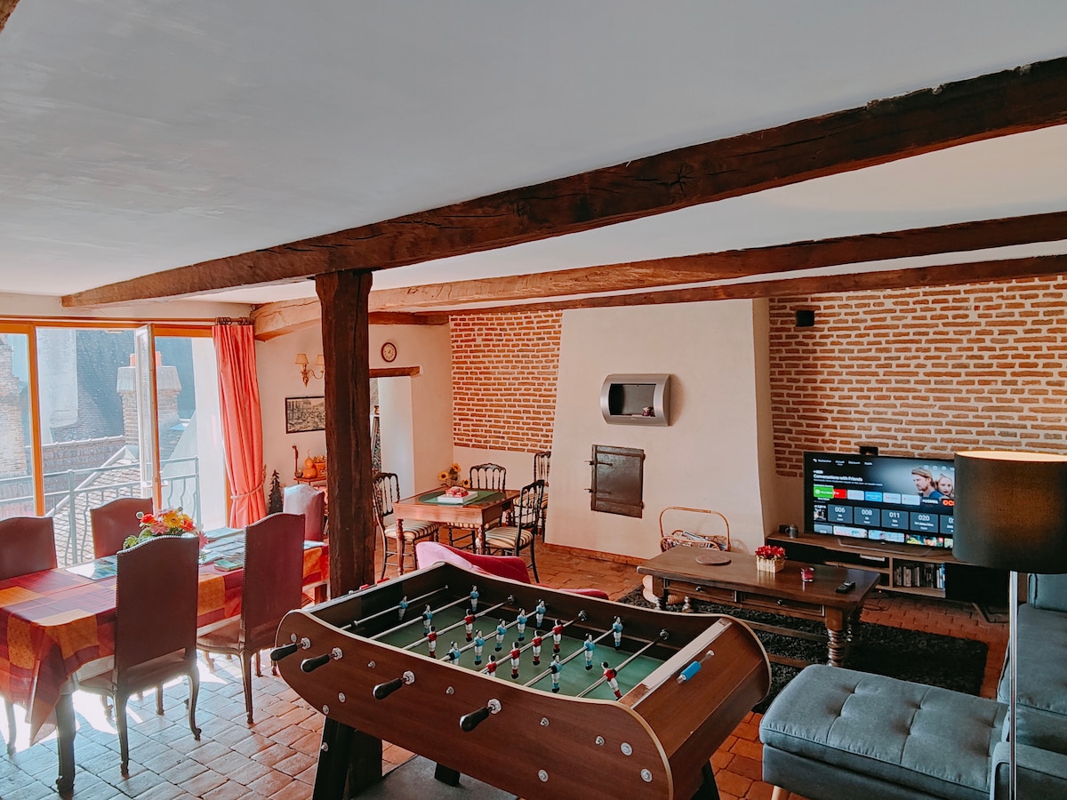 An expansive living area features a foosball table at the center, complemented by a dining table and comfortable seating options. Large windows allow natural light to fill the space, highlighting the exposed wooden beams and brick wall accents.