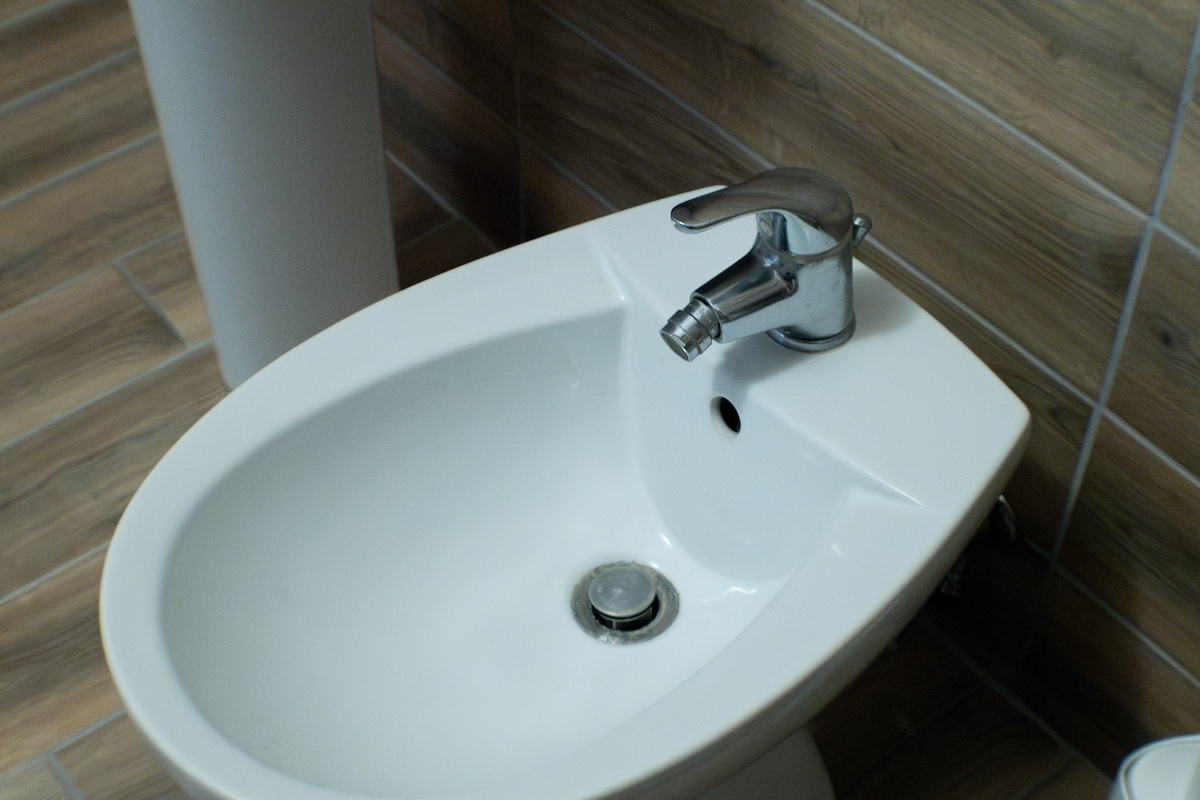 A clean, white bidet is showcased against a backdrop of textured wooden tiles. The shiny chrome faucet stands out, and the drainage outlet is visible at the base. Soft, neutral lighting enhances the space, providing a calm setting.