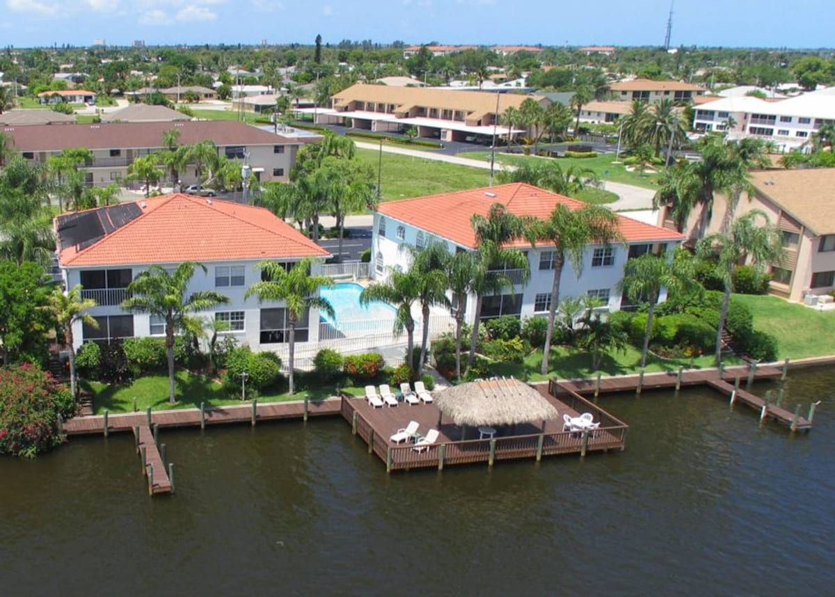 Aerial view of the waterfront property shows two connected buildings with red-tiled roofs. A large pool is visible within a landscaped area, surrounded by palm trees. A wooden deck with a Tiki Hut is positioned by the water, offering outdoor seating options. Neighboring homes are seen in the background.