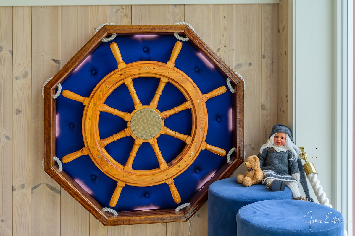 A colorful wooden ship's wheel is mounted on a blue background, framed by a dark wooden border. Near the wall, a plush doll and a teddy bear are positioned on a round blue stool, adding a cozy touch to the nautical decor.