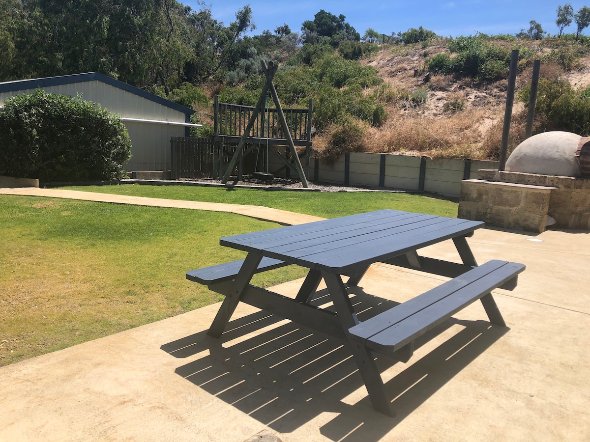 An outdoor dining area features a dark wooden picnic table positioned on a paved surface. The surrounding yard includes a swing and climbing frame on the lawn, with a stone pizza oven visible in the background. The area is bordered by lush greenery.