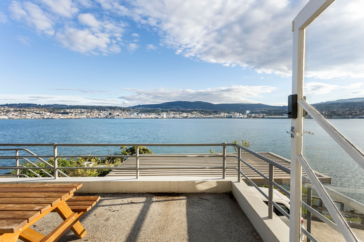 A spacious deck is visible, featuring wooden seating and railings that provide an unobstructed view of the tranquil water. The distant city skyline and rolling hills create a serene backdrop under a partly cloudy sky.