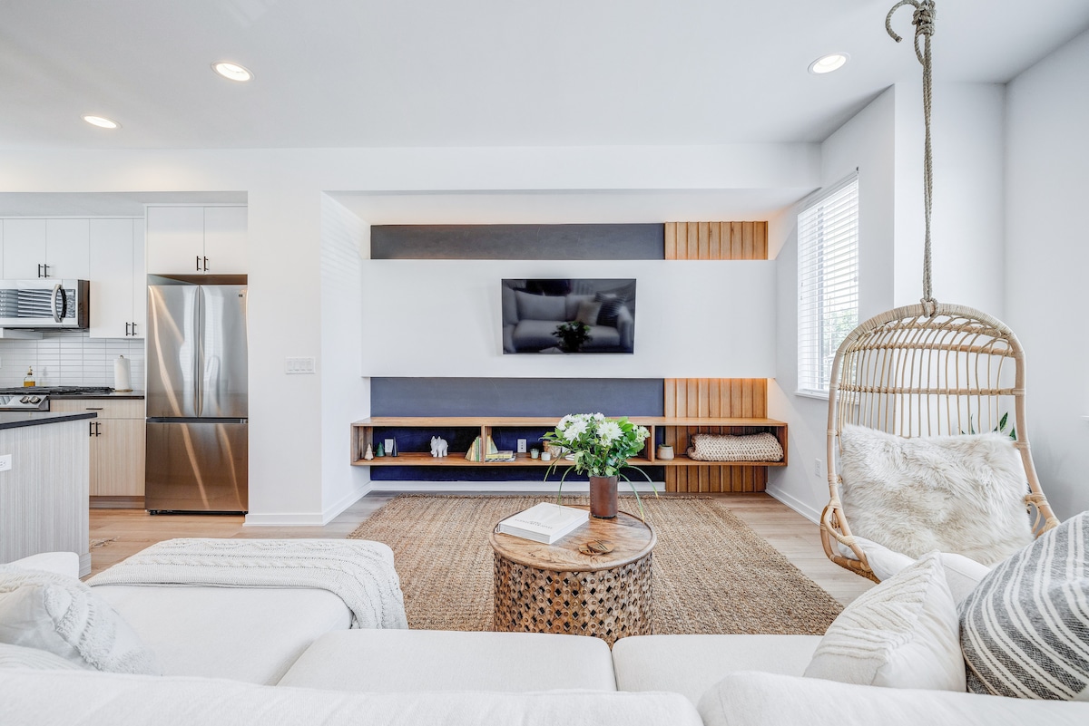A modern living area is presented with a neutral color palette, featuring a comfortable sectional sofa and a decorative natural fiber rug. An accent chair swings gently in the corner, and an entertainment unit houses a flat-screen television. Light enters through the nearby window, enhancing the space.