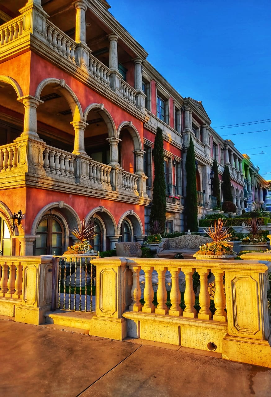 A grand building showcases classic architectural elements with arched windows and ornate columns. Vibrant coral tones adorn the facade, complemented by neatly trimmed greenery and decorative plants. The structure is framed by a stylized stone fence, creating an inviting entrance along the walkway.