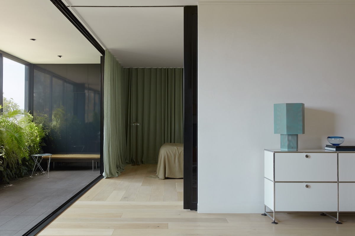A modern entryway reveals a spacious living area with large glass doors that invite natural light. Delicate green curtains frame a cozy sleeping area in the background. A white cabinet and a turquoise lamp add subtle color to the minimalist design, complementing the hardwood flooring.