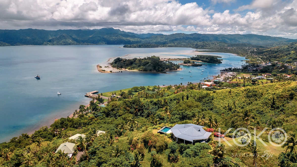 A panoramic view captures the serene Savusavu Bay, showcasing sandy beaches and lush green hills. The coastline is dotted with boats and the vibrant town is visible in the distance, framed by mountainous terrain and partially cloudy skies.